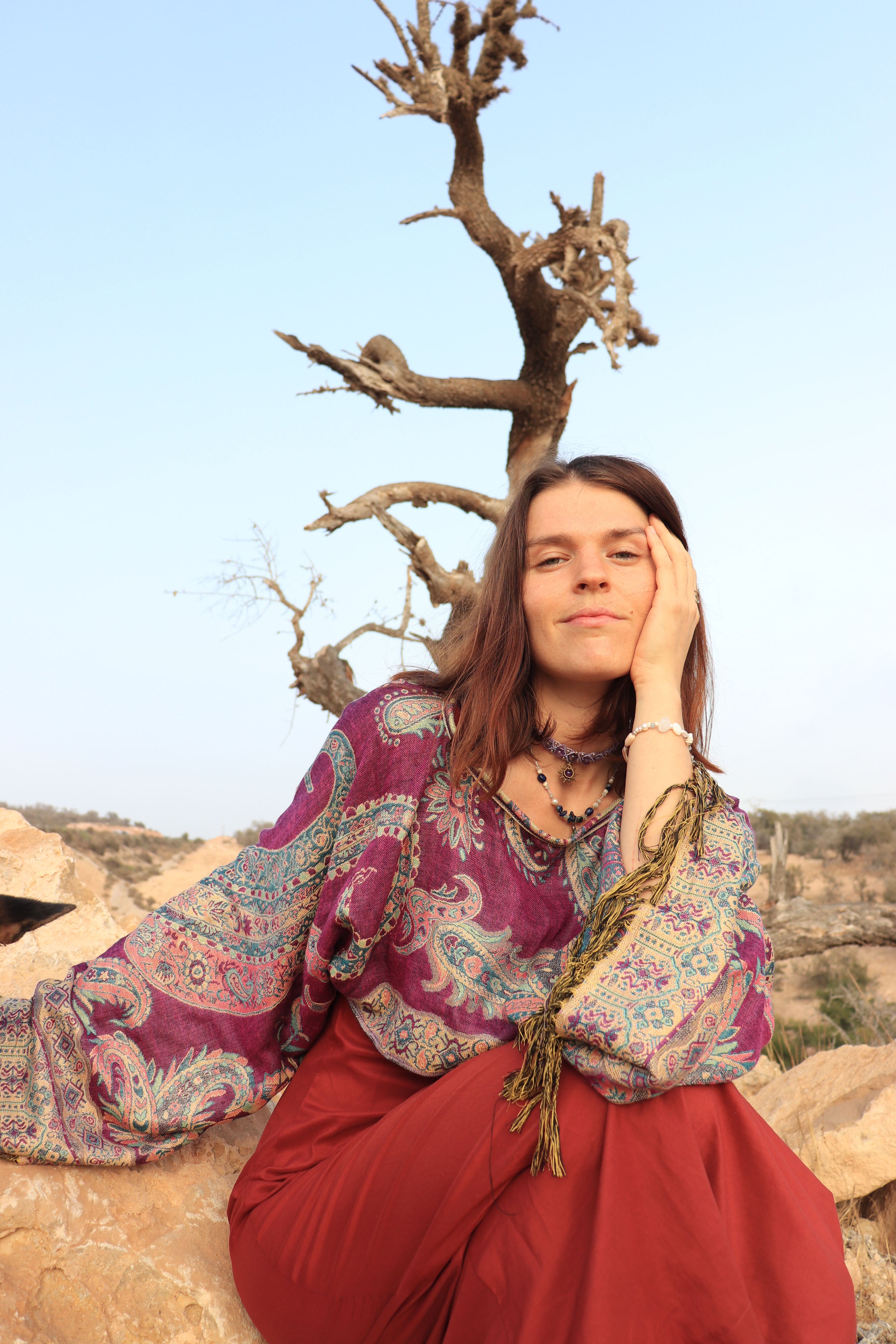 Woman in a patterned top and red skirt sitting on rocks with a tree in the background