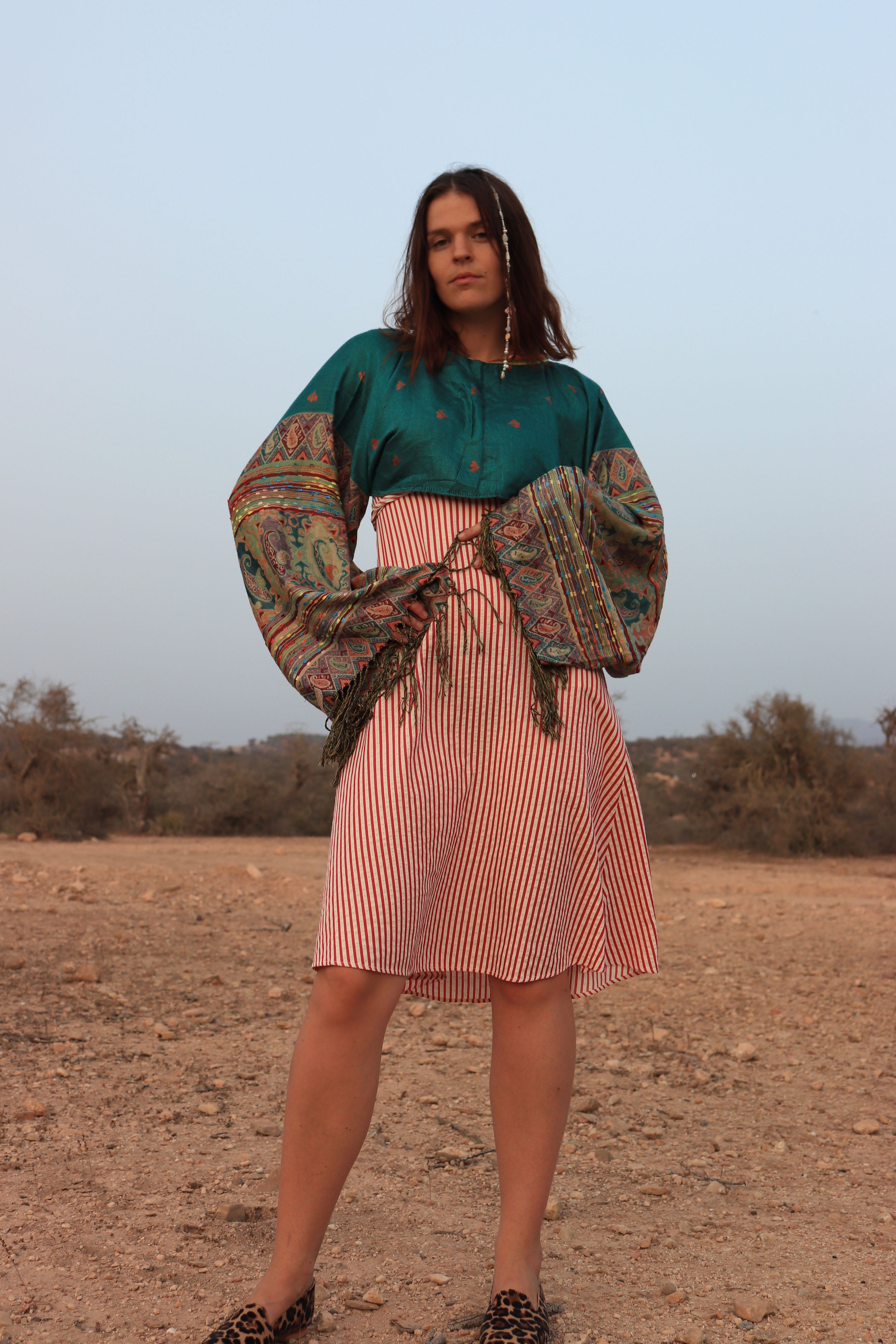Woman in a green top and pink skirt with a patterned jacket standing in a desert-like setting.