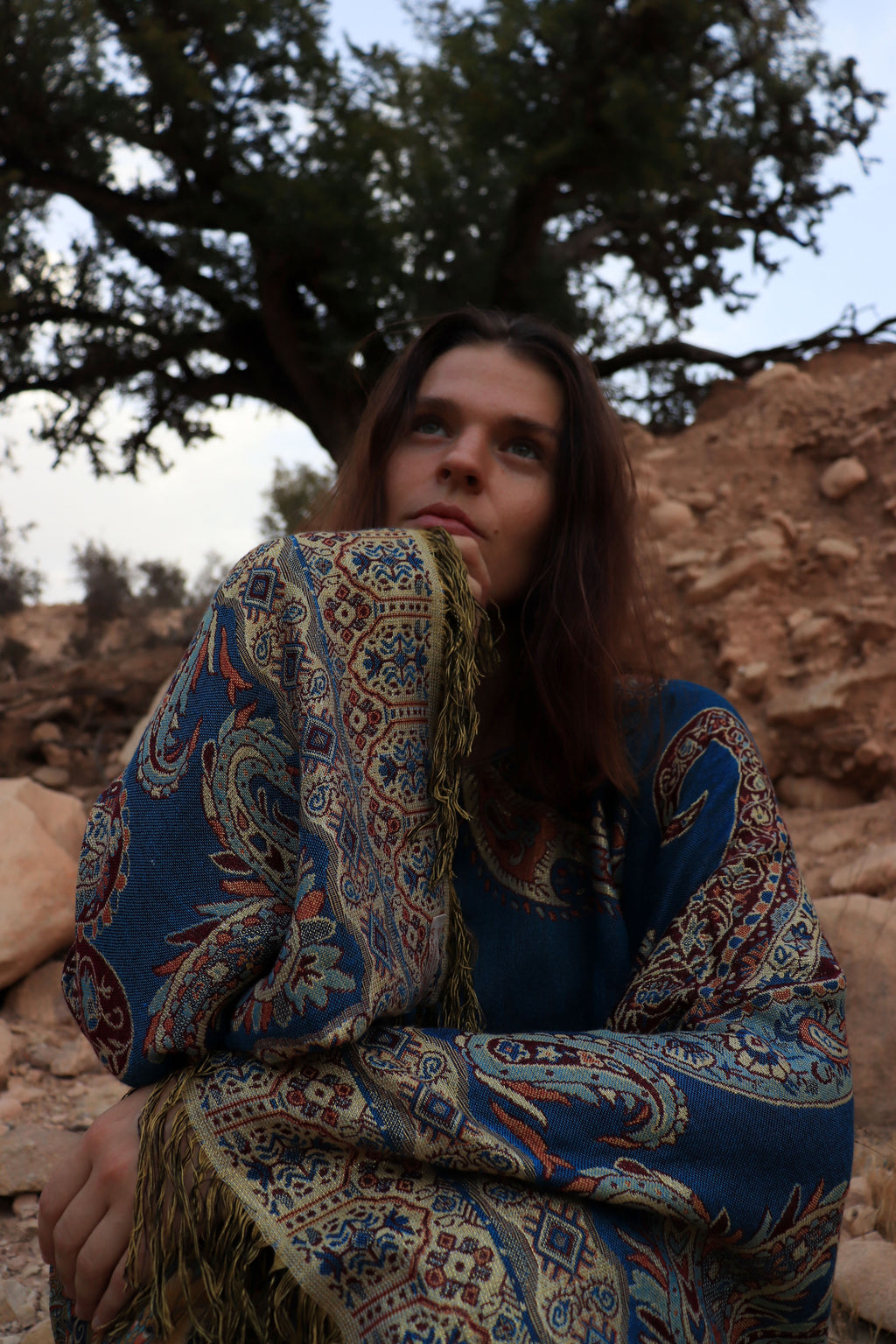 Woman wrapped in a patterned shawl sitting under a tree with rocky terrain.