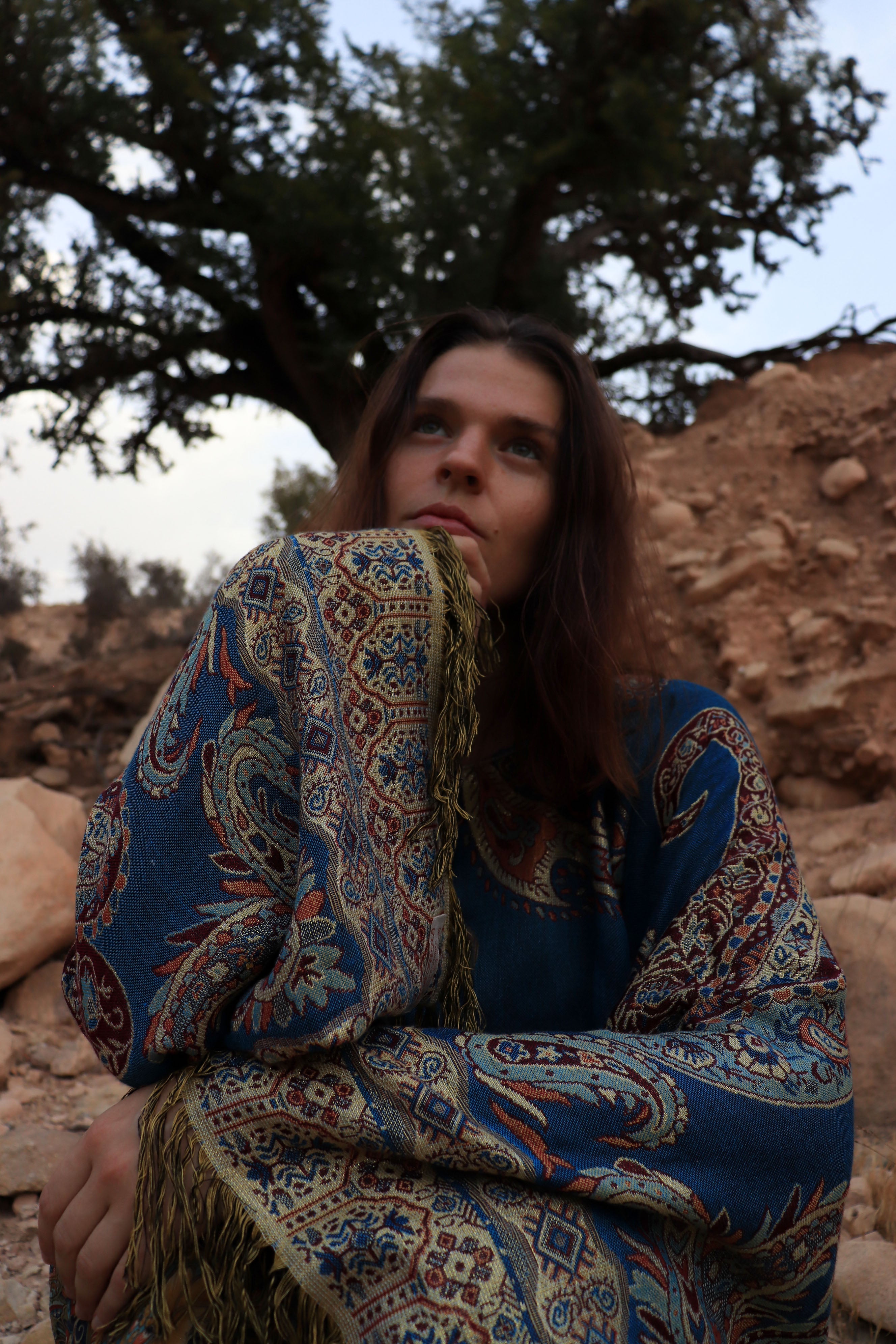 Woman wrapped in a patterned shawl sitting under a tree with rocky terrain.